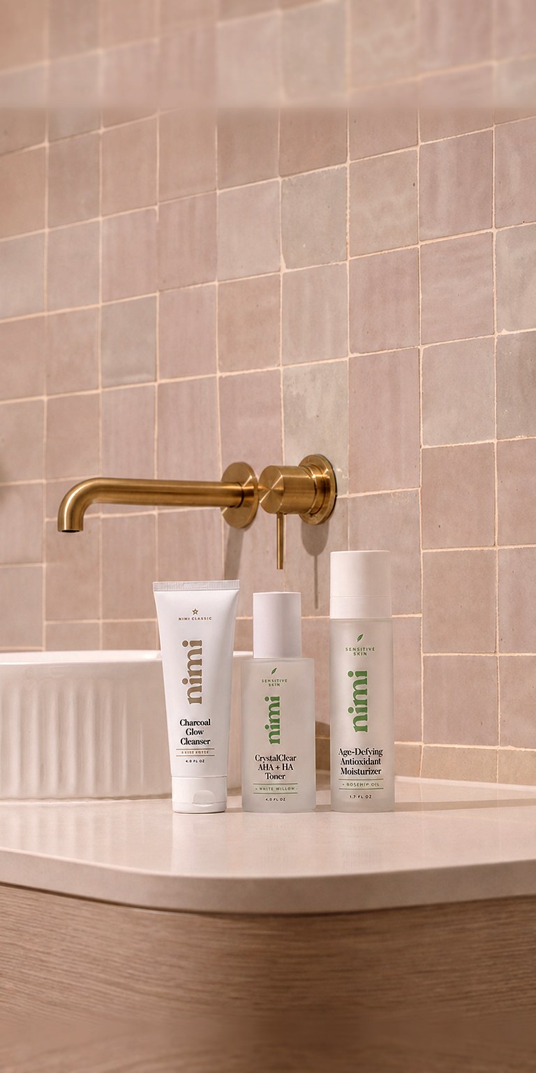 Nimi Charcoal Glow Cleanser, CrystalClear AHA + HA Toner, and Age-Defying Antioxidant Moisturizer on a bathroom vanity beneath a brushed-gold tap