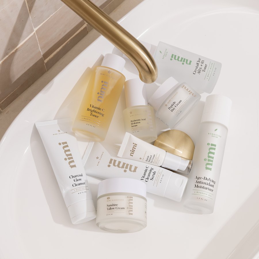 Nimi bottles, jars, and tubes gathered in a white basin beneath a brushed gold faucet, including Charcoal Glow Cleanser, Vitamin C Brightening Toner, and Sunshine Tallow Cream